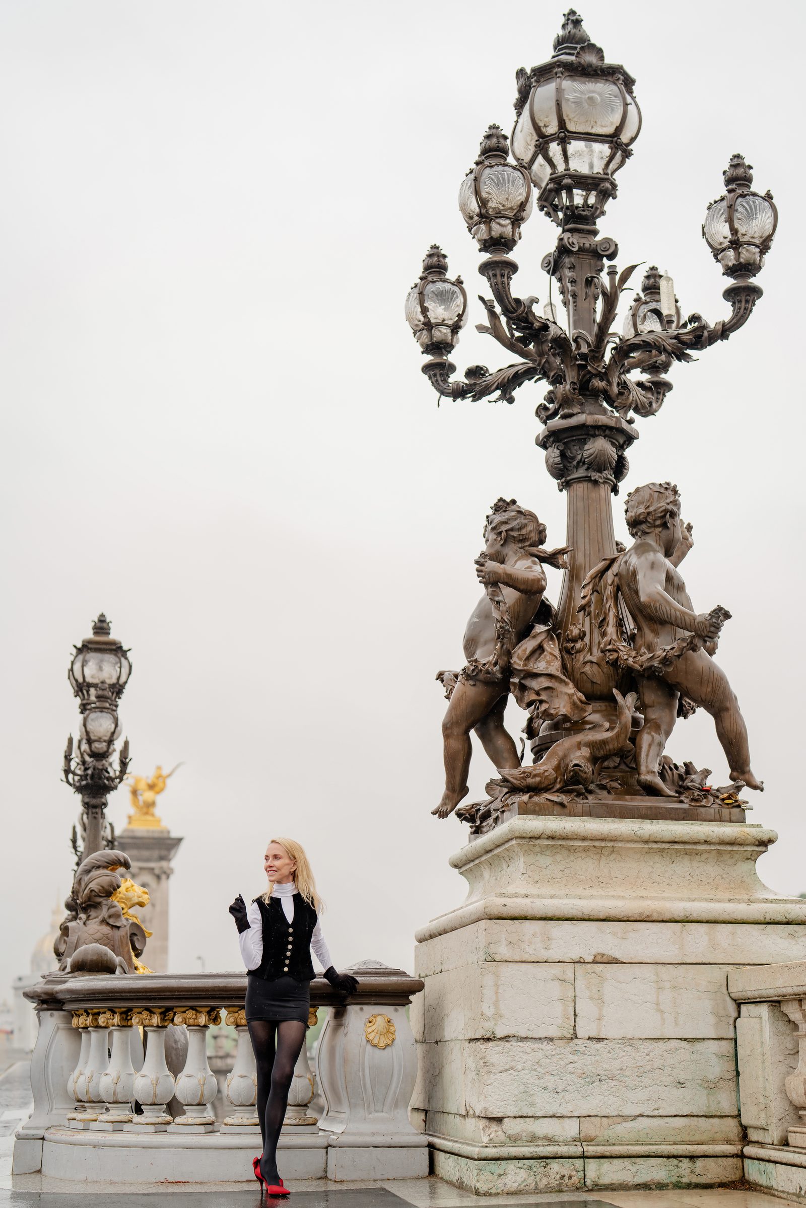 Lise Liddell on the Pont Alexandre III in Paris, in a black vest, white blouse and red shoes, beneath a bronze lamp post.